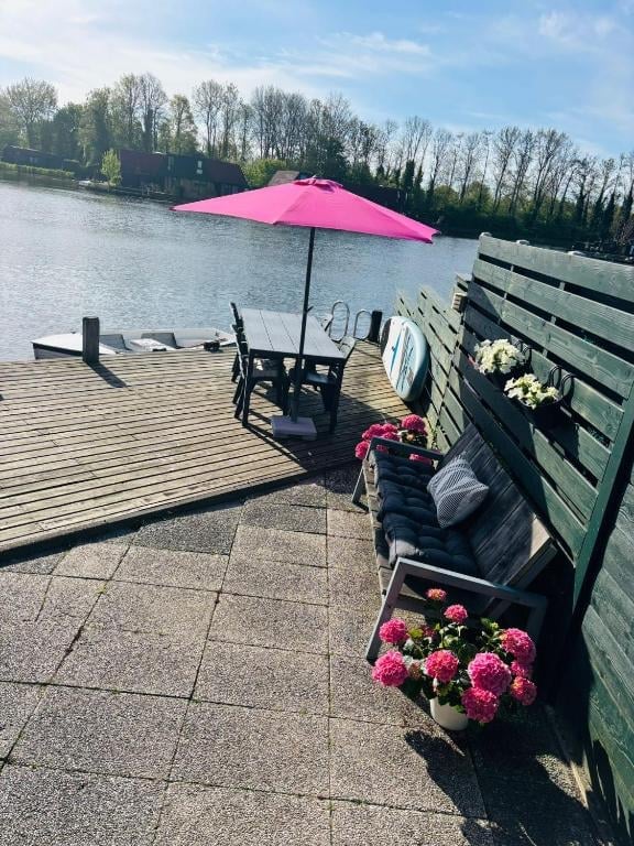Waterside patio with pink umbrella shading black chairs overlooking a lake, bordered by pink flowers and wooden fence