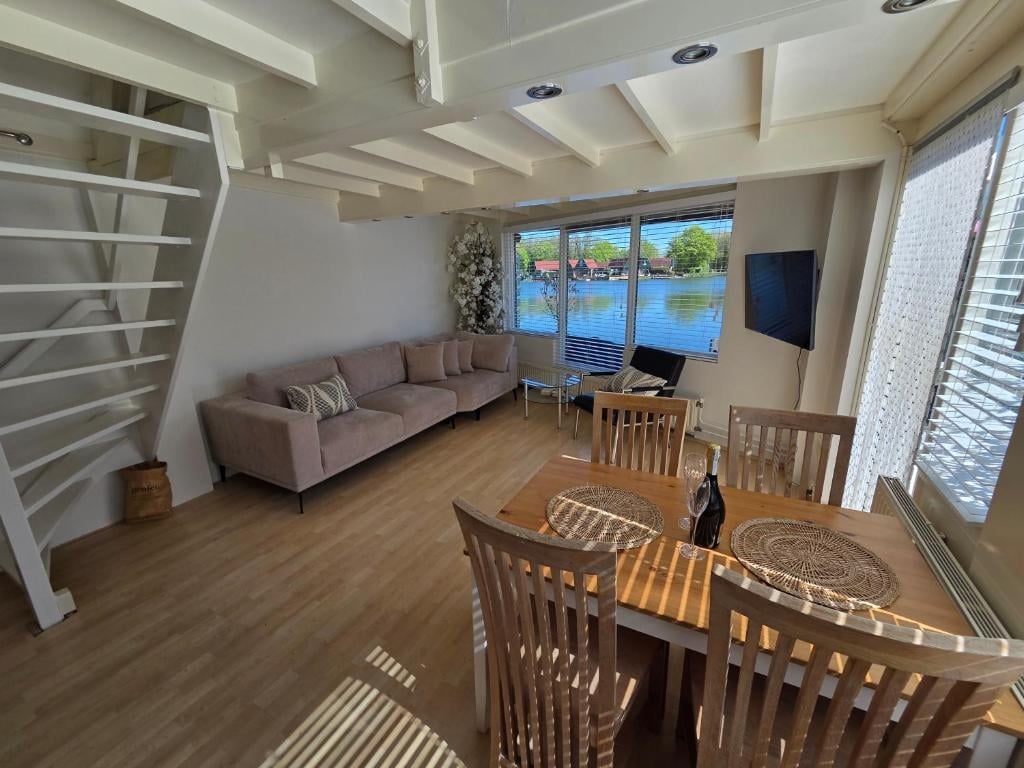 Modern open-concept living room with wooden dining table, gray sofa, large windows, and exposed ceiling beams with natural light