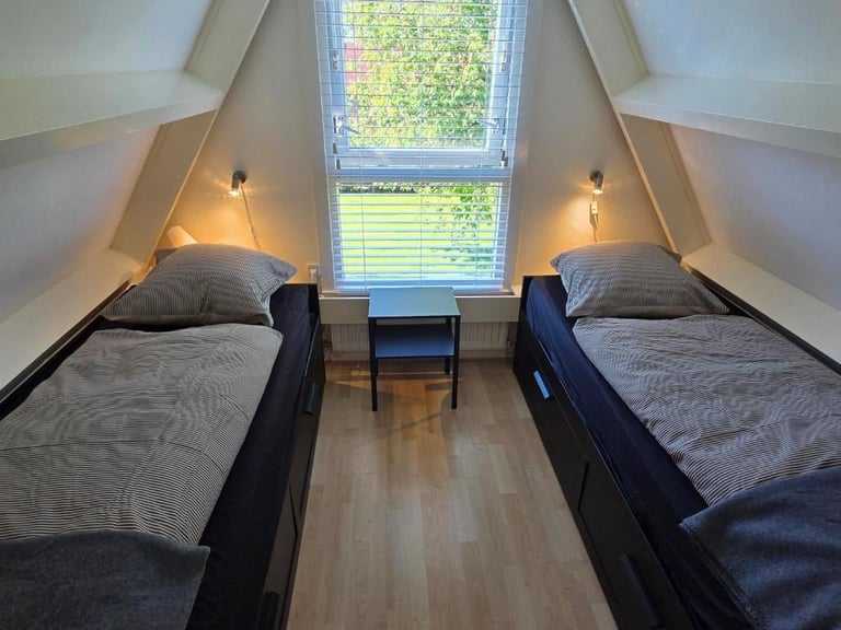 Cozy attic bedroom with two twin beds, warm bedside lighting, and a large window overlooking greenery